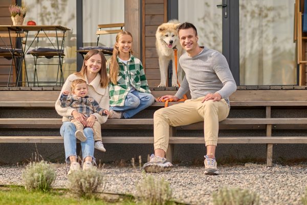 Good day. Young adult man with dog woman with child and school-age girl smiling looking at camera sitting on steps of country house on fine afternoon